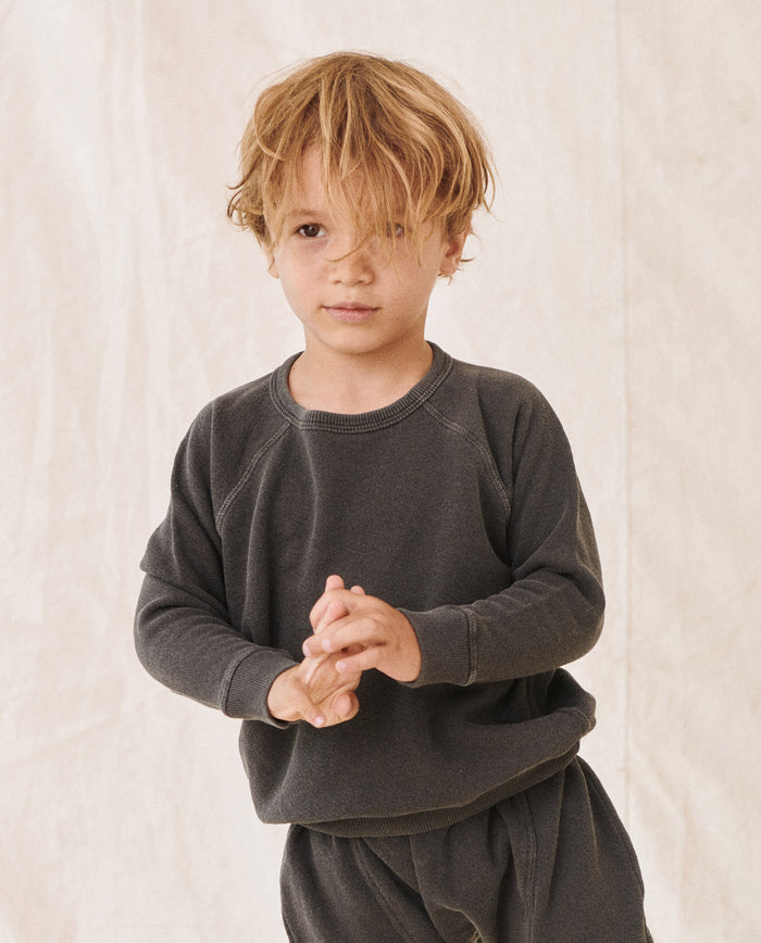 Against a plain background, a young child with tousled blonde hair stands with hands clasped together, dressed in a dark long-sleeve shirt and matching pants, and styled in The Little College Sweatshirt in Washed Black from THE GREAT. HOL 21 LITTLE.