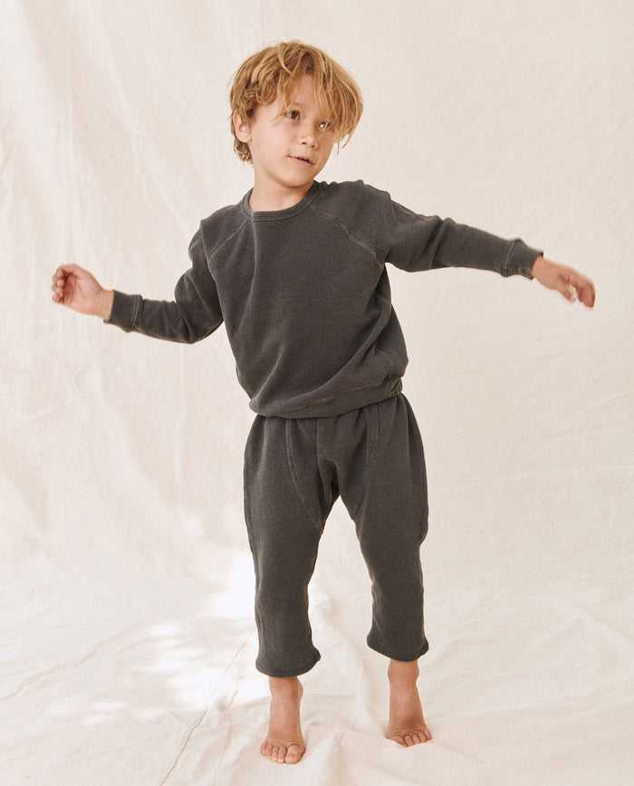 A young child with wavy hair stands barefoot on a white fabric backdrop, wearing a washed black "The Little College Sweatshirt" by THE GREAT. HOL 21 LITTLE and matching pants, with arms slightly outstretched.