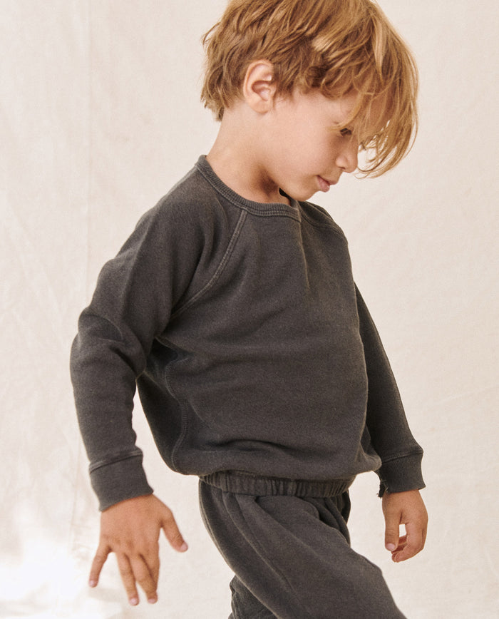 A young child with light brown hair wearing The Little College Sweatshirt in washed black by THE GREAT. HOL 21 LITTLE stands against a beige background while looking down.