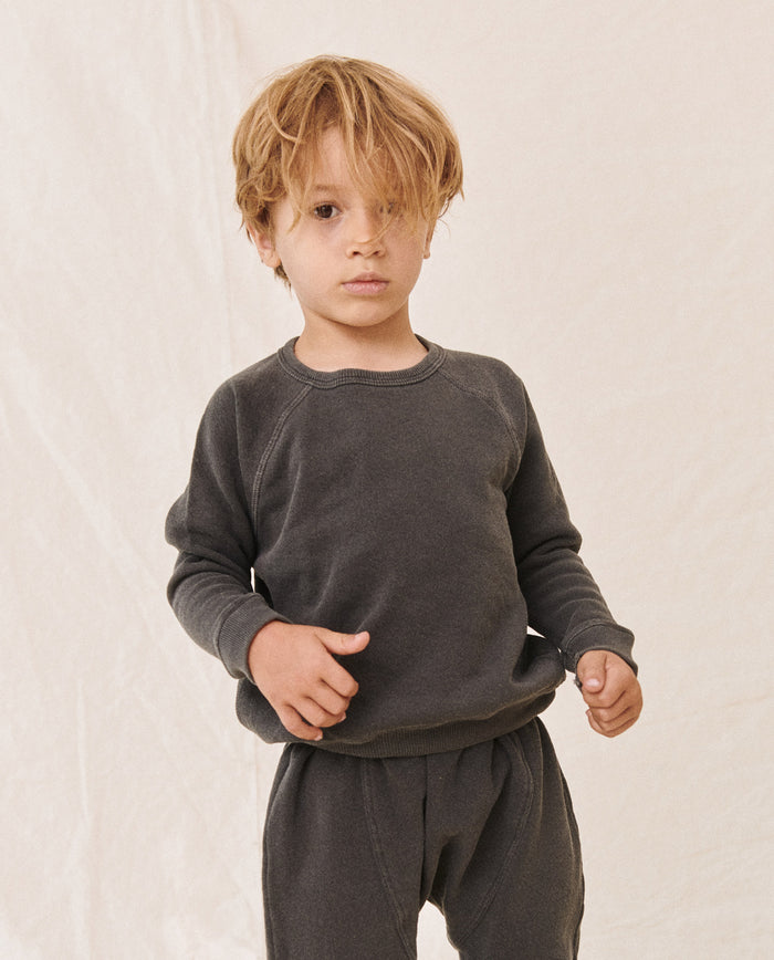 A young child stands against a plain backdrop, wearing THE GREAT. HOL 21 LITTLE's The Little College Sweatshirt in washed black and matching pants. Their hair is slightly tousled, covering part of their forehead.