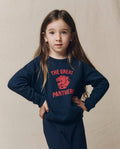 A young child with long brown hair stands with hands on hips, wearing THE GREAT. F26 D1 The Little College Sweatshirt w/ Growling Panther in True Navy—a soft French Terry piece featuring “The Great Panthers” and a bold red panther graphic—against a plain beige background.