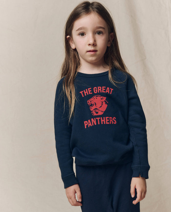 A young child with long brown hair stands against a beige backdrop, wearing THE GREAT.’s The Little College Sweatshirt w/ Growling Panther in True Navy, featuring soft French Terry and “The Great Panthers” in red text.
