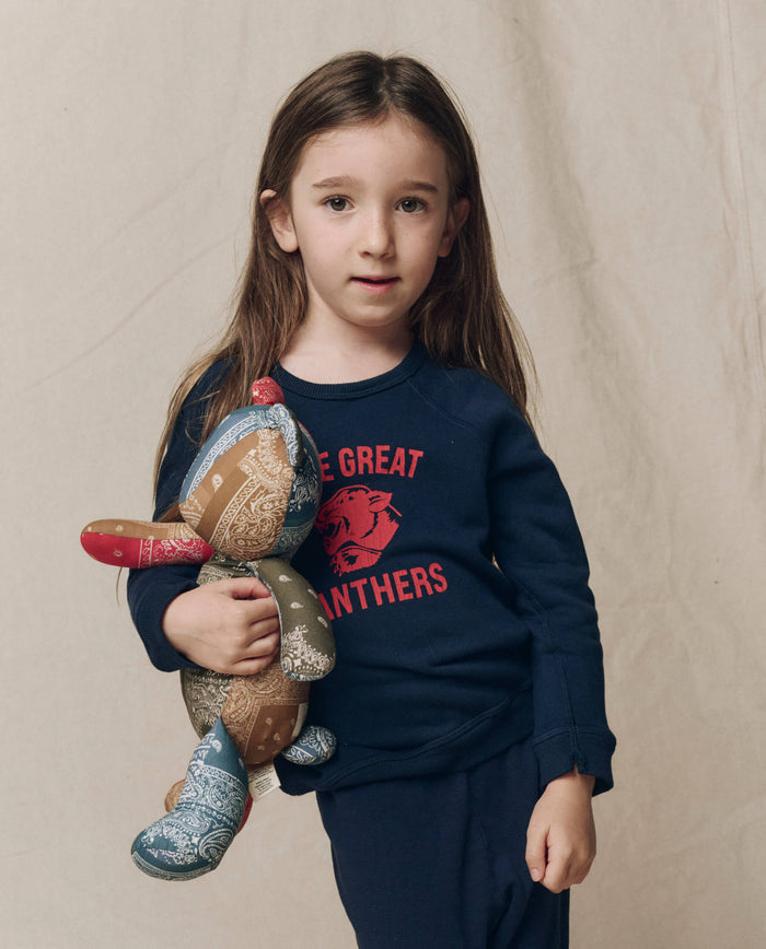 A young girl with long brown hair wears THE GREAT. F26 D1 The Little College Sweatshirt w/ Growling Panther in True Navy, paired with matching pants and holding a patchwork stuffed animal, standing against a neutral background.