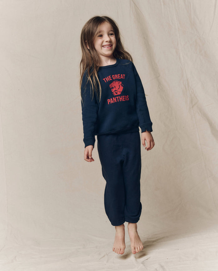 A young child wearing THE GREAT. F26 D1 The Little College Sweatshirt with Growling Panther in True Navy is jumping in front of a plain beige backdrop.