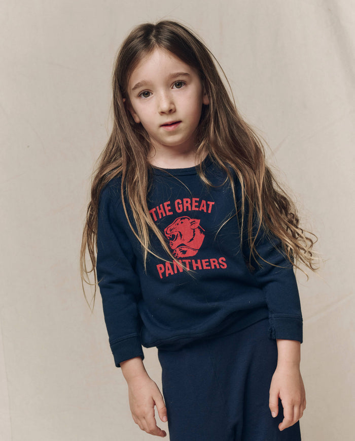 A young child with long brown hair wears THE GREAT. F26 D1’s The Little College Sweatshirt w/ Growling Panther in True Navy and matching soft French Terry pants, standing against a plain light background.