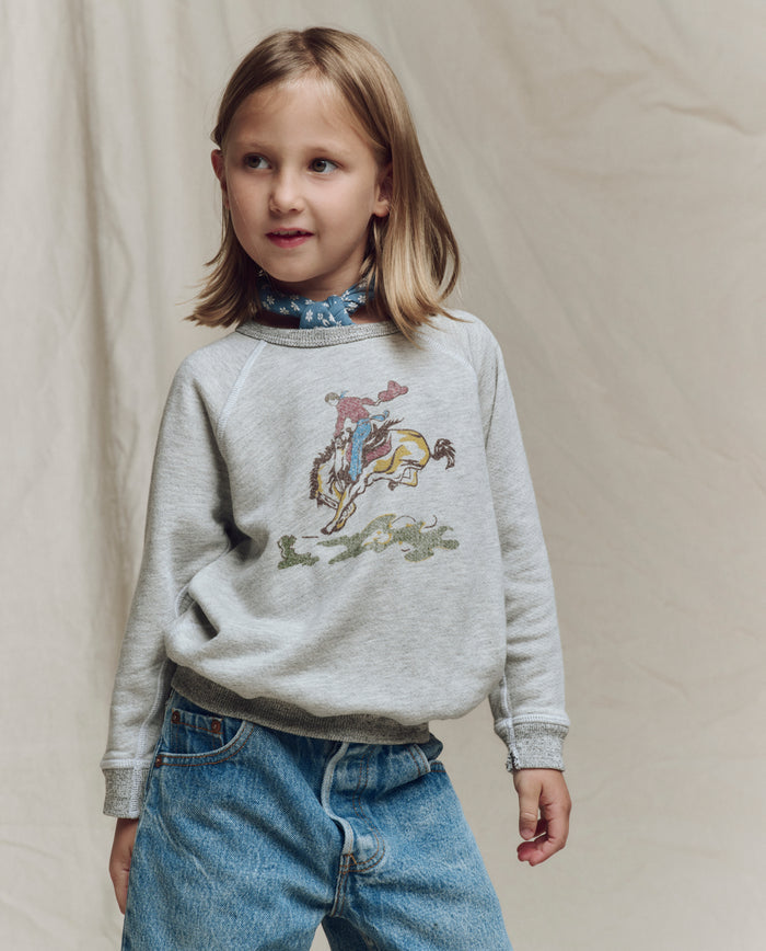 A young child wears THE GREAT. SP26 LITTLES The Little College Sweatshirt w/ Rider Graphic in soft heather grey, paired with blue jeans, standing before a neutral fabric backdrop.