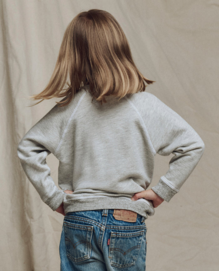 A child with shoulder-length light brown hair, hands on hips, faces away in THE GREAT. SP26 LITTLES "The Little College Sweatshirt w/ Rider Graphic" in Soft Heather Grey and blue jeans against a neutral background.