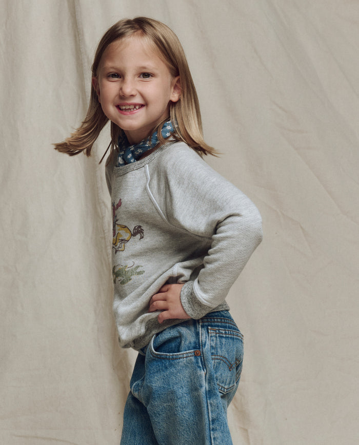A young girl with shoulder-length blonde hair models THE GREAT. SP26 LITTLES’ The Little College Sweatshirt w/ Rider Graphic in Soft Heather Grey and blue jeans, smiling sideways with one hand on her hip against a plain beige background.