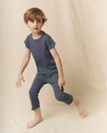 A young boy with brown hair poses barefoot in front of a beige fabric backdrop, wearing THE GREAT. LITTLE’s The Little Jersey Crop in Solid Washed Navy—a handcrafted t-shirt and matching jersey crop pant set featuring an elastic waist, made with care in L.A.