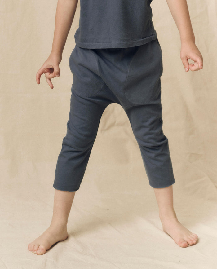 A child stands barefoot on a beige surface, wearing THE GREAT. LITTLE's The Little Jersey Crop in Washed Navy—a blue shirt paired with matching elastic-waist blue pants, arms gently bent and fingers relaxed.
