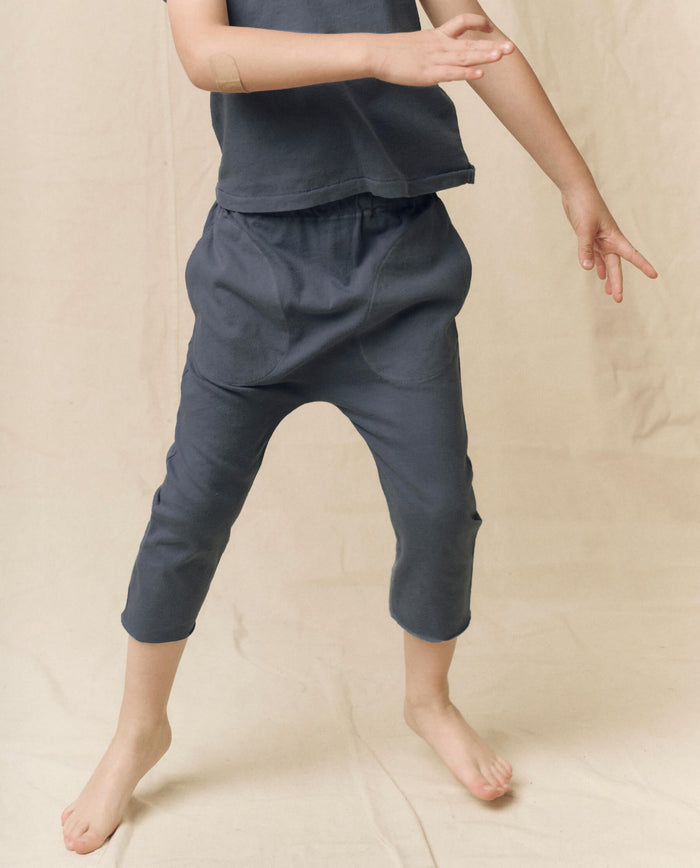 A child wearing THE GREAT. LITTLE’s The Little Jersey Crop Solid in Washed Navy—loose-fitting crop pants with an elastic waist and a coordinating top—stands barefoot on a beige surface, one arm raised and one leg slightly lifted in motion. Each piece is thoughtfully handcrafted in L.A.
