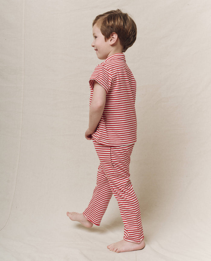 A child wearing The Little Lacrosse Polo in Sun Cherry Stripe by THE GREAT. SP25 POLO LOUNGE LITTLE stands sideways against a light background, exuding a playful sportswear vibe.