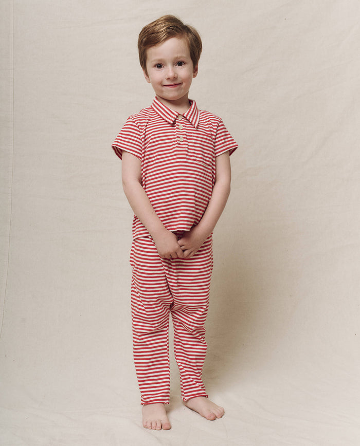 A child stands barefoot against a plain background, wearing The Little Lacrosse Polo in Sun Cherry Stripe by THE GREAT. SP25 POLO LOUNGE LITTLE, smiling with the enthusiasm of a lacrosse player ready to take on the game.