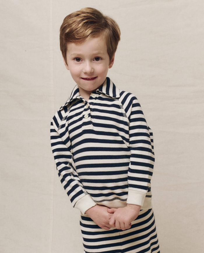 A child wearing The Little Rugby Sweatshirt in Navy Sailor Stripe by THE GREAT. SP25 POLO LOUNGE LITTLE stands against a plain background, looking towards the camera.
