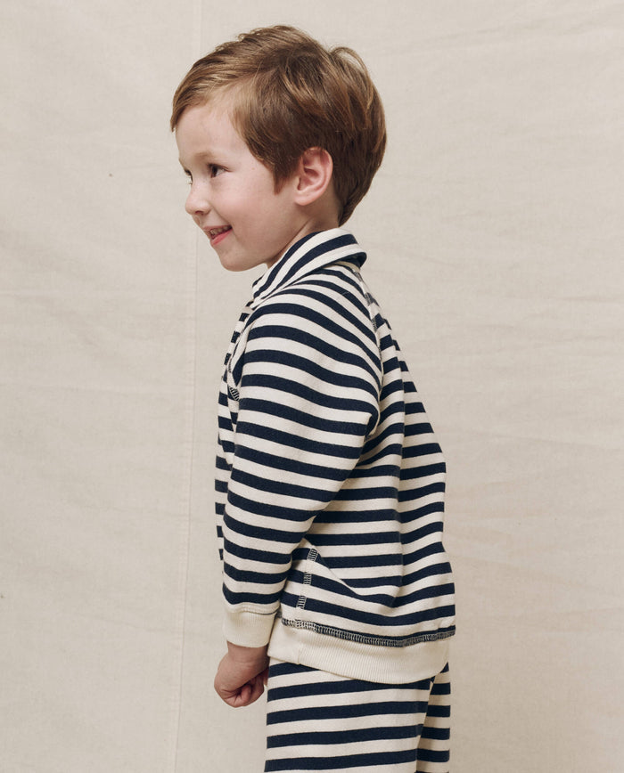 A child wearing The Little Rugby Sweatshirt in Navy Sailor Stripe from THE GREAT. SP25 POLO LOUNGE LITTLE, stands sideways against a plain background, capturing playful innocence.