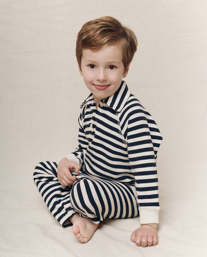 A child sits cross-legged on a light background, slightly smiling while wearing The Great. SP25 Polo Lounge Little's "The Little Rugby Sweatshirt" in Navy Sailor Stripe.