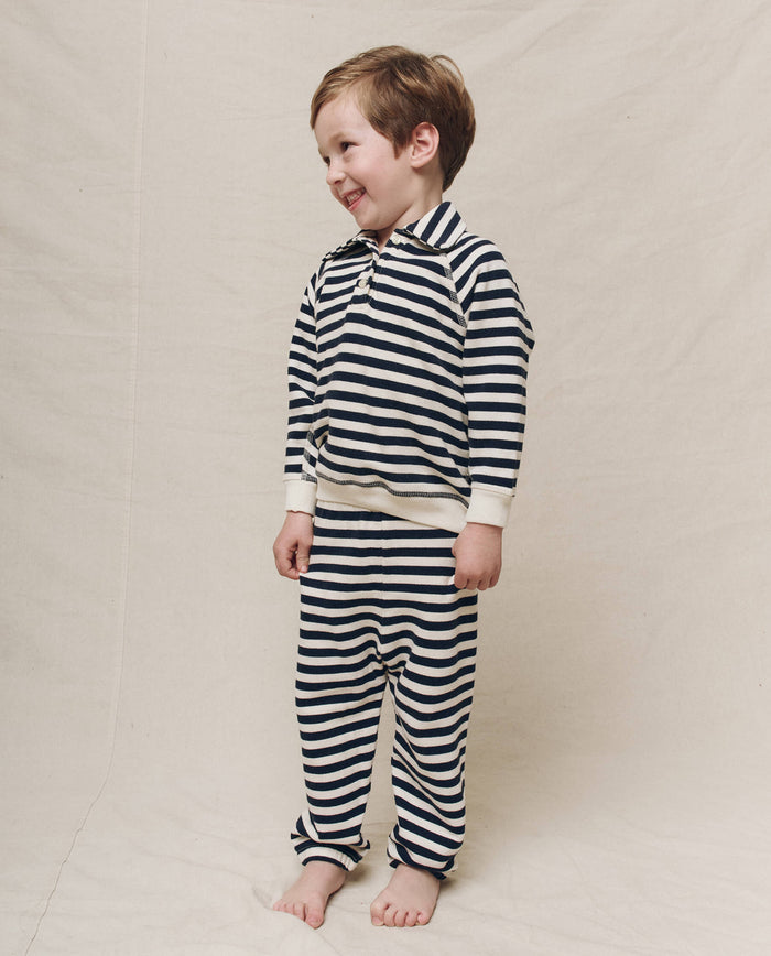 A child in The Little Rugby Sweatshirt by THE GREAT. SP25 POLO LOUNGE LITTLE, featuring a Navy Sailor Stripe pattern, smiles against a plain background.