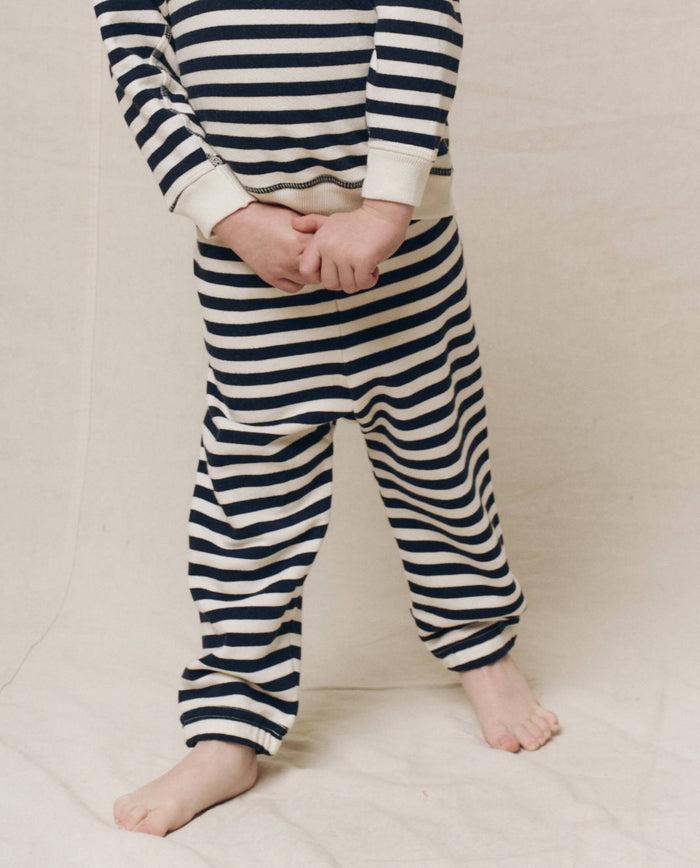 A child in the navy sailor stripe Little Stadium Sweatpant by THE GREAT. stands barefoot on the beige floor, hands clasped in front.