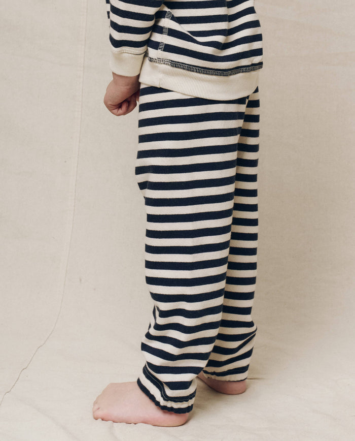 A child in navy sailor striped pajamas from THE GREAT.'s The Little Stadium Sweatpant collection stands barefoot on a neutral surface, arms at their sides.