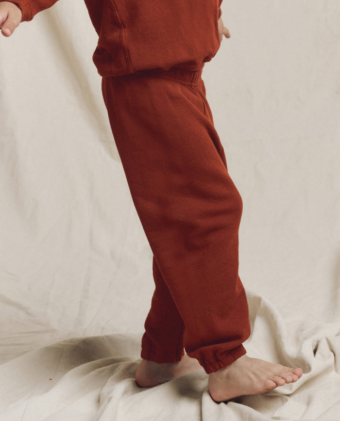 A person in THE GREAT. F25 LITTLES The Little Stadium Sweatpant, Solid -- Brick, and a matching sweatshirt stands barefoot on a light fabric surface, highlighting vintage athletic wear style.