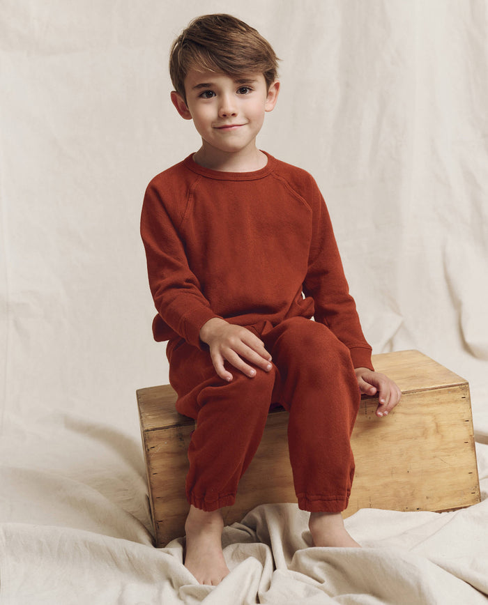 A young boy in THE GREAT. F25 LITTLES The Little Stadium Sweatpant, Solid -- Brick, and a matching long-sleeve top sits barefoot on a wooden box against a neutral fabric backdrop.