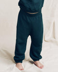 A child in THE GREAT. F25 LITTLES The Little Stadium Sweatpant, Solid -- Dark Slate, and a matching sweatshirt stands barefoot on a light fabric surface.
