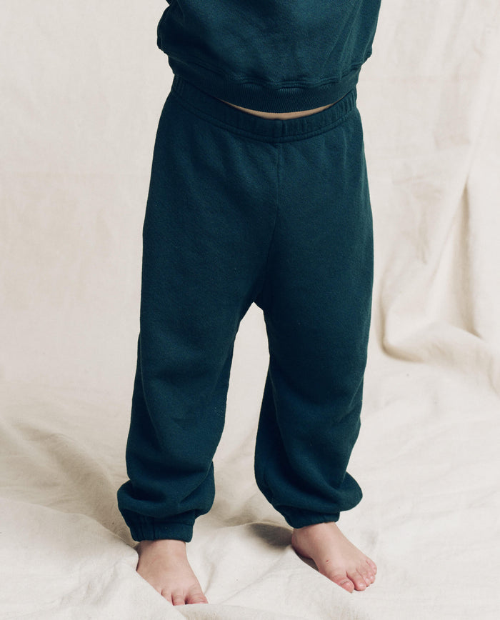 A child in THE GREAT. F25 LITTLES The Little Stadium Sweatpant, Solid -- Dark Slate, and a matching sweatshirt stands barefoot on a light fabric surface.