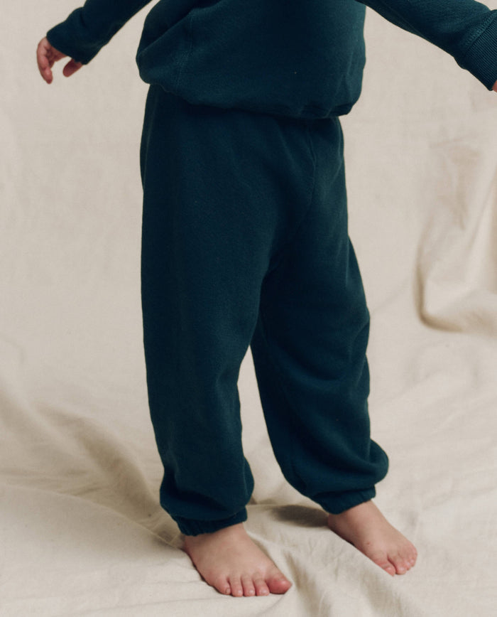 A young child wears THE GREAT. F25 LITTLES The Little Stadium Sweatpant, Solid -- Dark Slate, standing barefoot on a light fabric surface with only the lower body in view—a nod to L.A. crafted vintage athletic style.