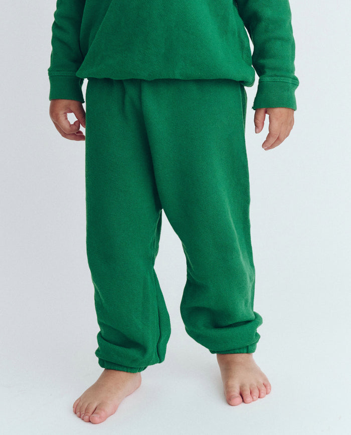 A person wearing a green long-sleeved shirt and THE GREAT. HOL 23 LITTLE's The Little Stadium Sweatpant in Solid -- Holly Leaf, standing barefoot against a plain white background. The photo shows the person from the shoulders down, giving off a vibe reminiscent of vintage athletic wear.