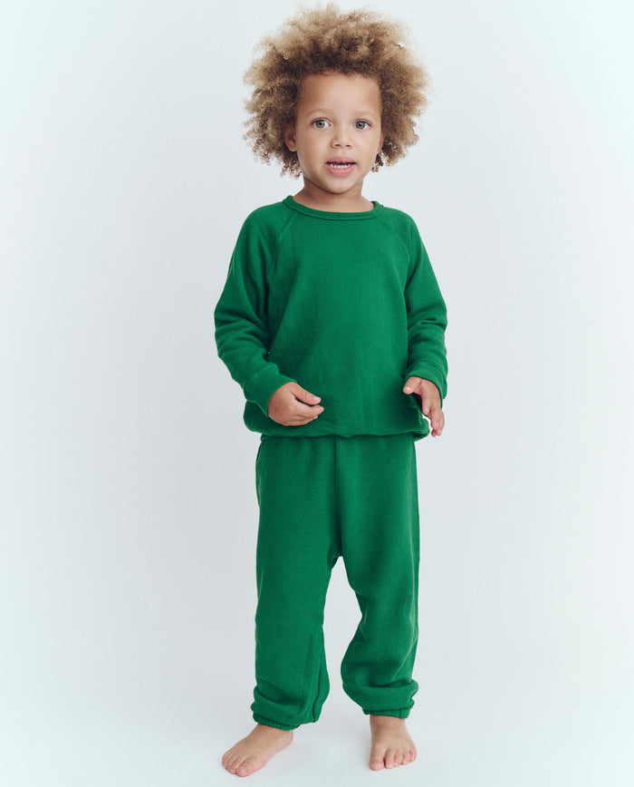 A child with curly hair stands barefoot against a white background, wearing a matching green long-sleeve shirt and The Little Stadium Sweatpant in Holly Leaf from THE GREAT. HOL 23 LITTLE, reminiscent of vintage athletic wear.