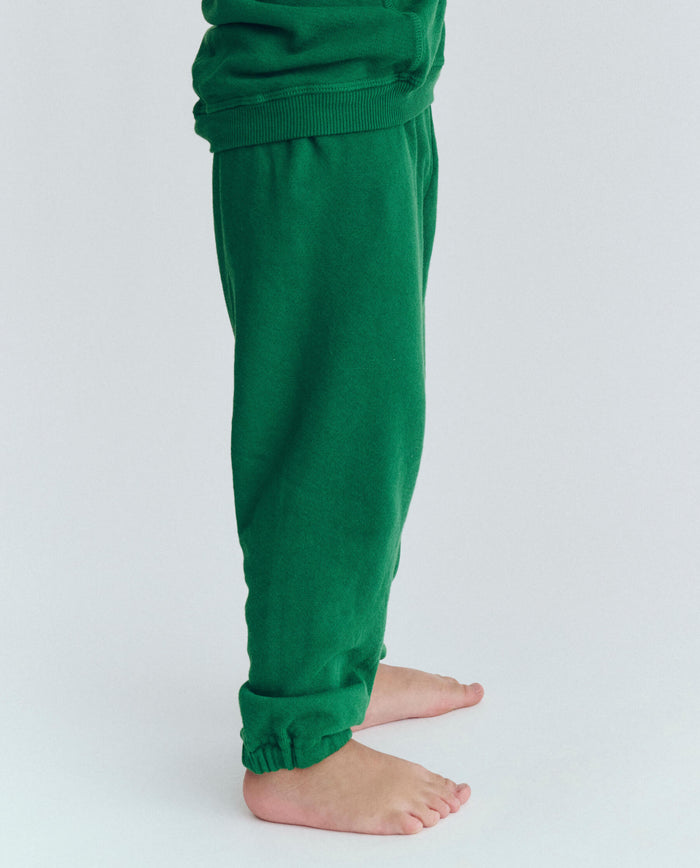 A person standing barefoot on the floor, wearing The Little Stadium Sweatpant in Holly Leaf by THE GREAT. HOL 23 LITTLE, with only their lower legs and feet visible.