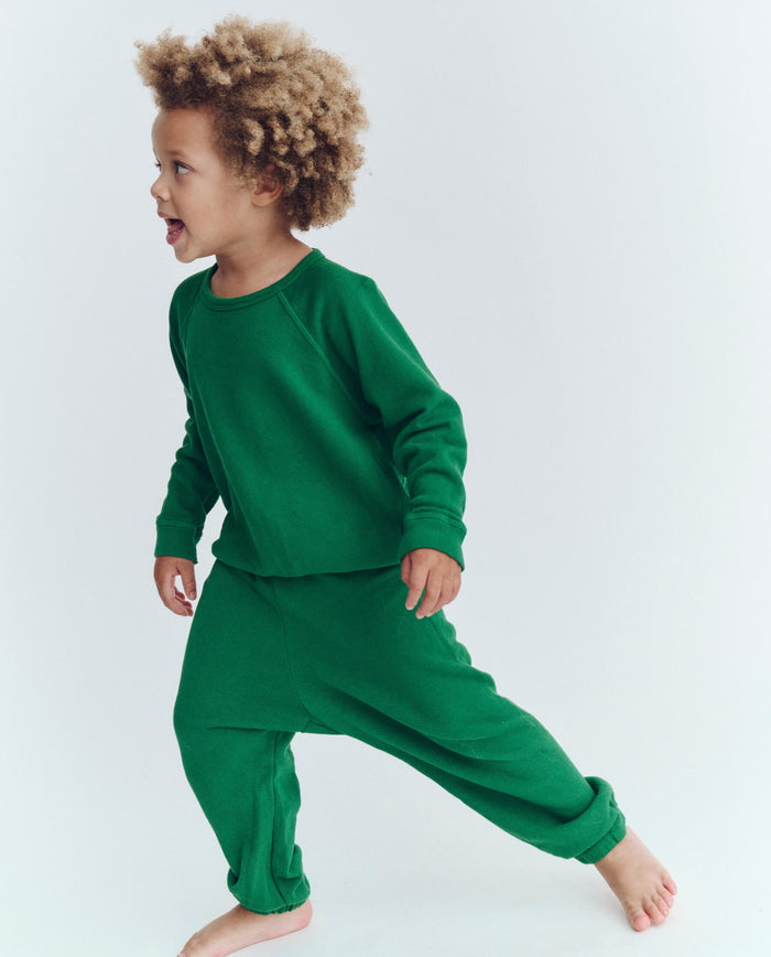 A young child with curly hair, wearing a green long-sleeve shirt and THE GREAT. HOL 23 LITTLE The Little Stadium Sweatpant in Holly Leaf, stands barefoot.