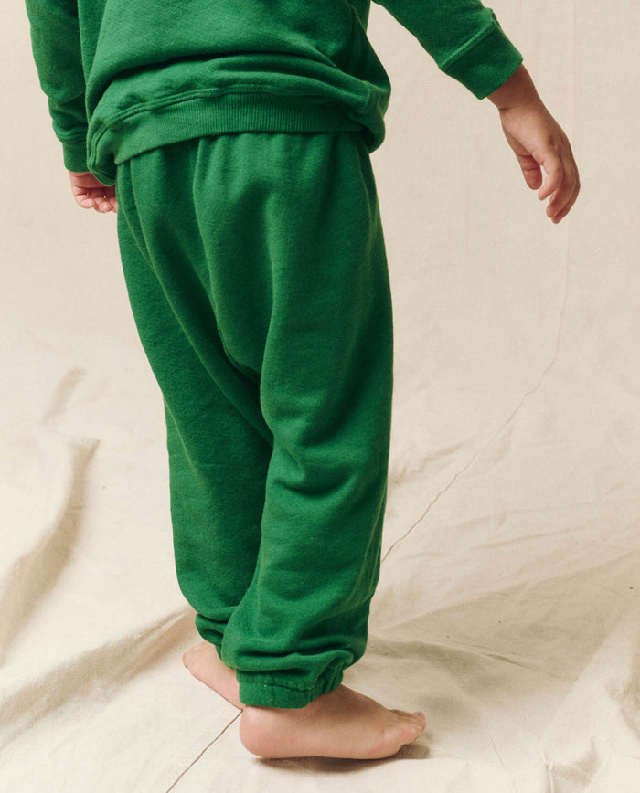 A child wearing The Little Stadium Sweatpant in Holly Leaf green from THE GREAT. HOL 23 LITTLE stands barefoot on a beige fabric surface, evoking the charm of vintage athletic wear.
