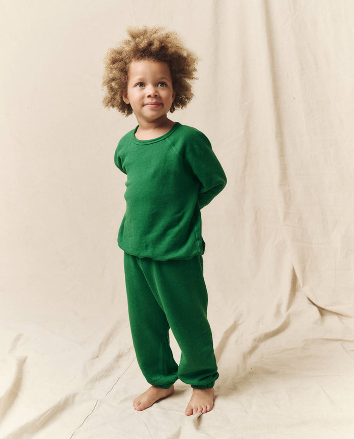 A child with curly hair stands barefoot against a beige background, showcasing handcrafted in Los Angeles vintage athletic wear with a green long-sleeve top and matching The Little Stadium Sweatpant in Holly Leaf by THE GREAT. HOL 23 LITTLE.
