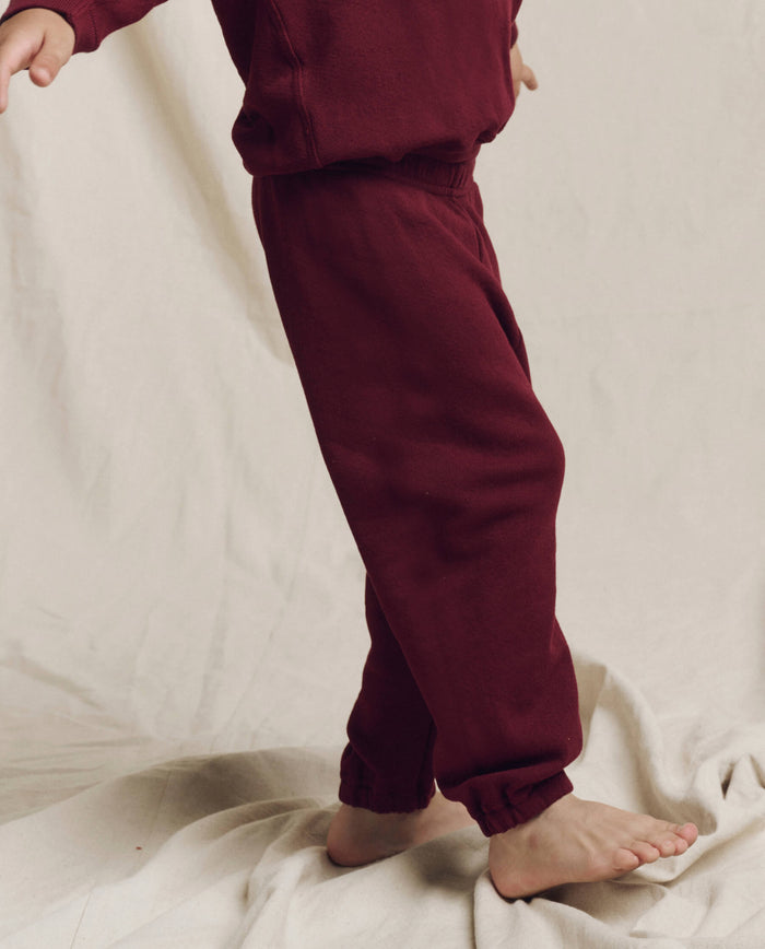 A child in a plum-colored tracksuit from The Little Stadium Sweatpant collection by THE GREAT. HOL 24 LITTLE stands barefoot on a cream-colored fabric backdrop.