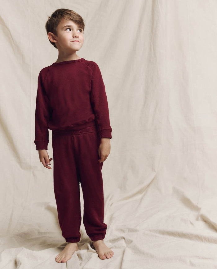A young boy in the plum Little Stadium Sweatpant by THE GREAT. HOL 24 LITTLE stands barefoot against a beige fabric backdrop, looking to his right for ultimate comfort sportswear.