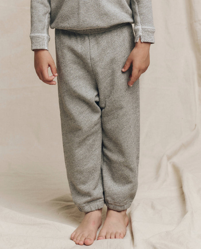 A child dressed in The Little Stadium Sweatpant, a solid Varsity Grey set from THE GREAT. LITTLE, stands barefoot on a light fabric background, evoking vintage athletic wear. Crafted in L.A., this set perfectly embodies a timeless blend of comfort and style.