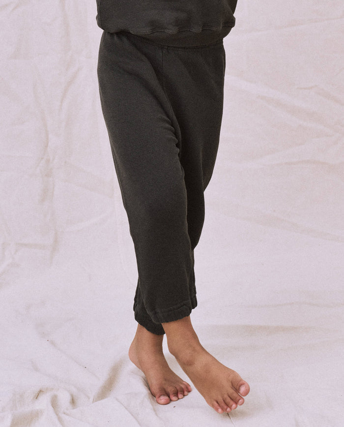 A child wearing The Little Stadium Sweatpant in Washed Black from THE GREAT. SP22 D2 LITTLE stands barefoot on a light fabric backdrop.
