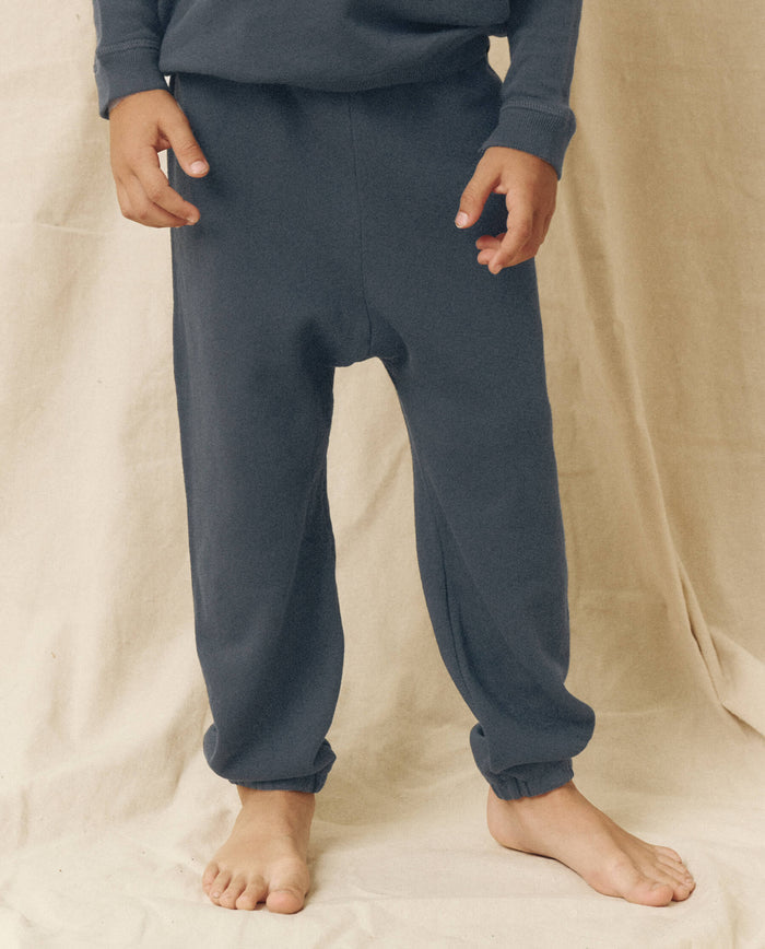 A child, barefoot on a beige fabric background, wears THE GREAT. FALL 22 LITTLE’s The Little Stadium Sweatpant in Solid -- Washed Navy with a matching top, capturing vintage athletic wear charm.