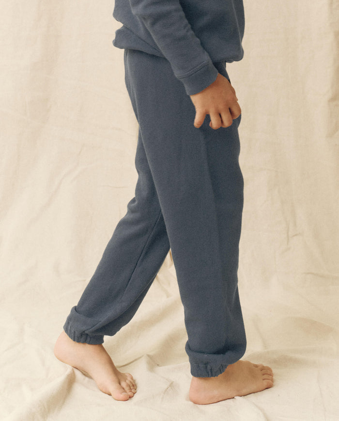 A child in THE GREAT. FALL 22 LITTLE The Little Stadium Sweatpant, Solid -- Washed Navy, stands barefoot on a cream fabric backdrop for a charming mommy & me moment.