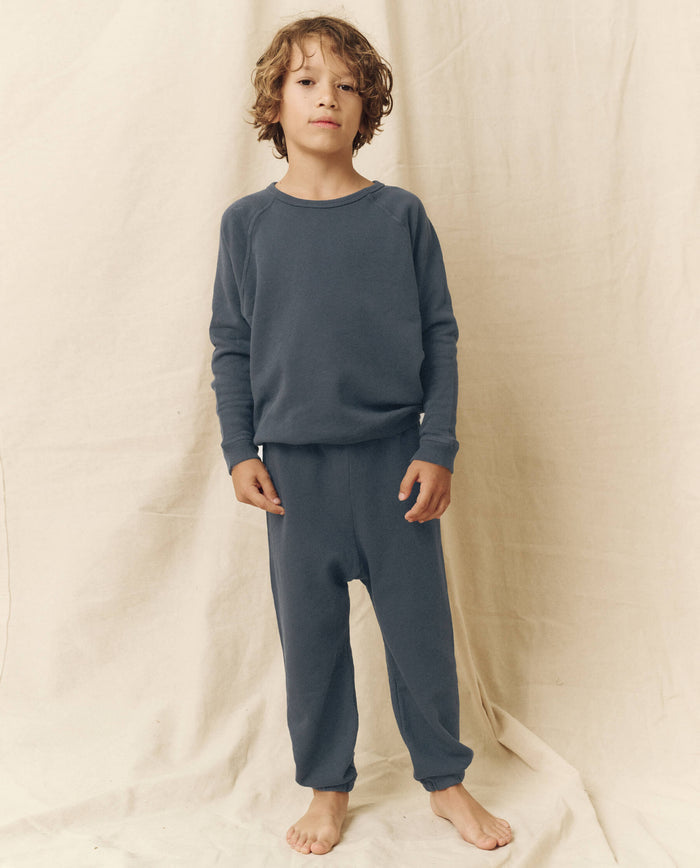 A young child with curly hair stands barefoot on a light fabric backdrop, wearing a blue long-sleeve top and THE GREAT. FALL 22 LITTLE’s The Little Stadium Sweatpant in Solid Washed Navy, inspired by vintage athletic wear.