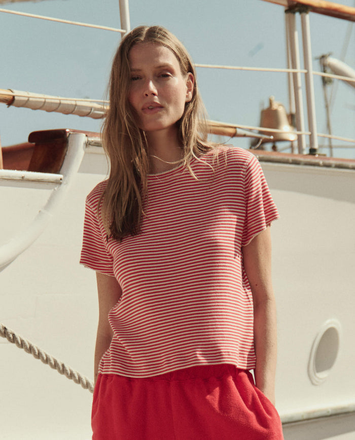 A woman wearing THE GREAT. SU26 The Little Tee in Pimento Stripe and red pants stands in front of a white boat on a sunny day.