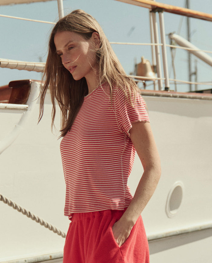 A woman in THE GREAT.'s The Little Tee -- Pimento Stripe stands in front of a yacht on a sunny day, paired with red shorts.