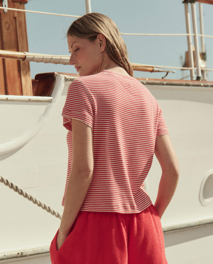 A person with light brown hair stands outdoors beside a white boat, wearing THE GREAT. SU26’s The Little Tee -- Pimento Stripe and red shorts.