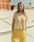 A woman stands outdoors in front of a white boat, wearing THE GREAT. SU26 The Little Tee. -- Soft Yellow Stripe and yellow drawstring pants, her hands in her pockets.