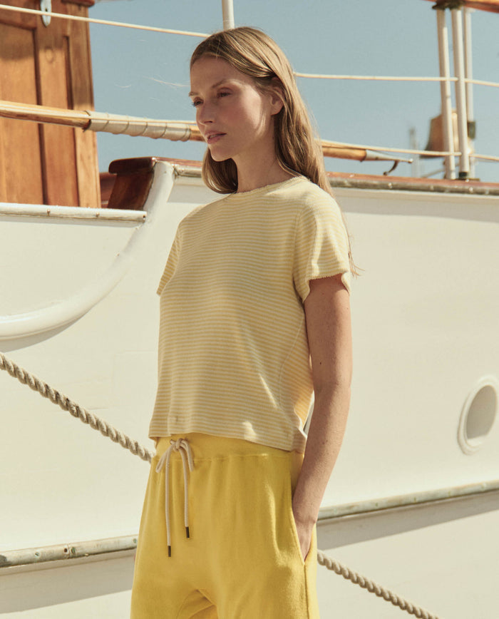 A woman wears THE GREAT. SU26's The Little Tee in Soft Yellow Stripe, paired with yellow pants, standing before a white boat on a sunny day.