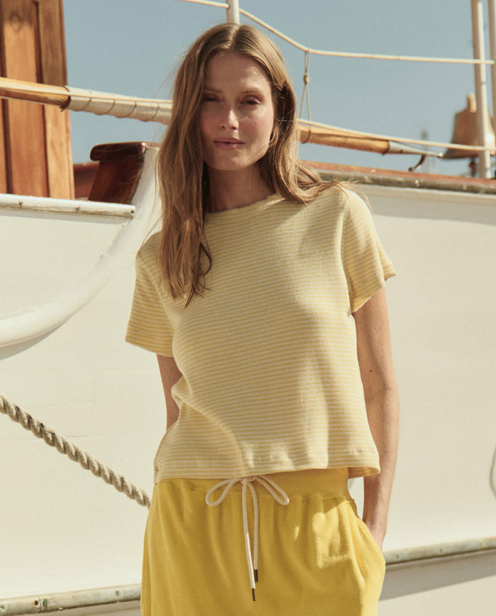 A woman wears THE GREAT. SU26 The Little Tee in Soft Yellow Stripe and yellow drawstring shorts while standing in front of a boat.