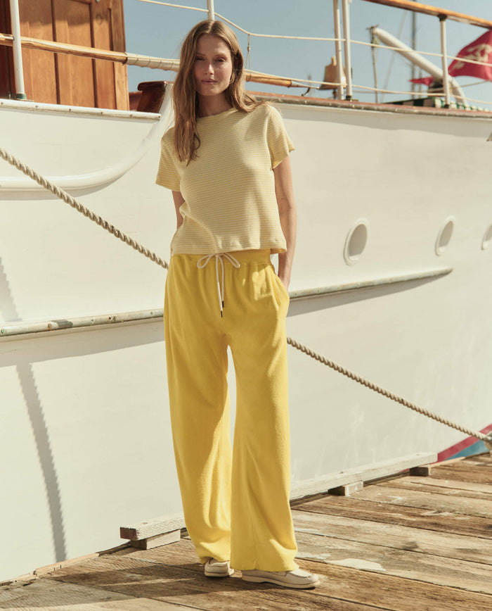 Woman stands on a dock before a white boat, wearing THE GREAT. SU26's The Little Tee in Soft Yellow Stripe, loose yellow pants, and beige shoes.