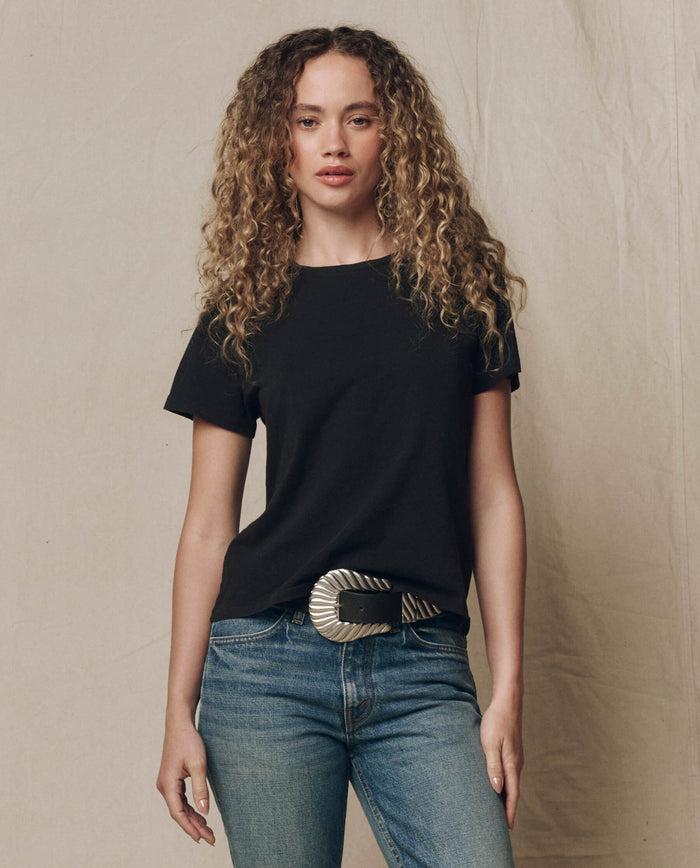 A person with long curly hair wears THE GREAT.'s The Little Tee. Solid -- Almost Black, blue jeans, and a wide silver belt, standing against a neutral background.