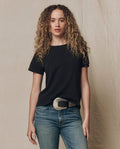 A person with long curly hair wears THE GREAT.'s The Little Tee. Solid -- Almost Black, blue jeans, and a wide silver belt, standing against a neutral background.